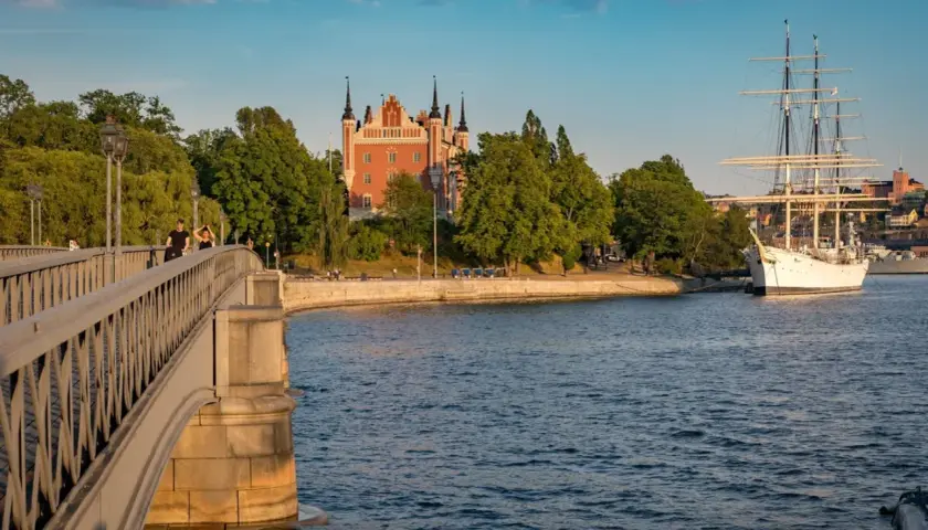 En vacker scen i Stockholm med en bro i förgrunden, ett rött tornliknande hus och ett stort vitt segelfartyg i vattnet.