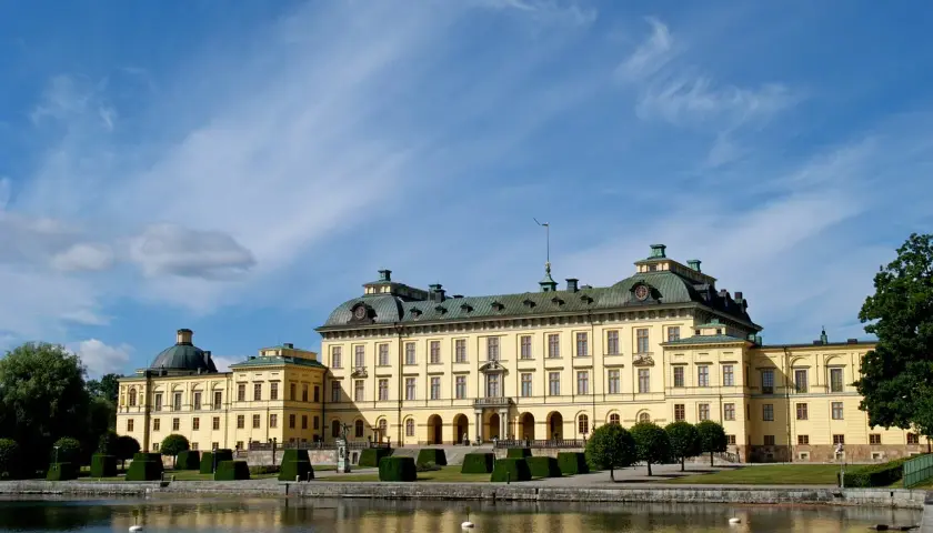 Drottningholm slott, ett gult barockpalats med gröna tak, speglar sig i vattnet under en blå himmel med tunna moln.