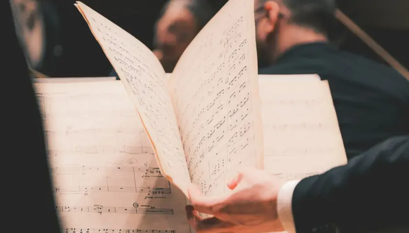 En hand bläddrar i noter under en lunchkonsert på operan. Musiker i bakgrunden förbereder sig.