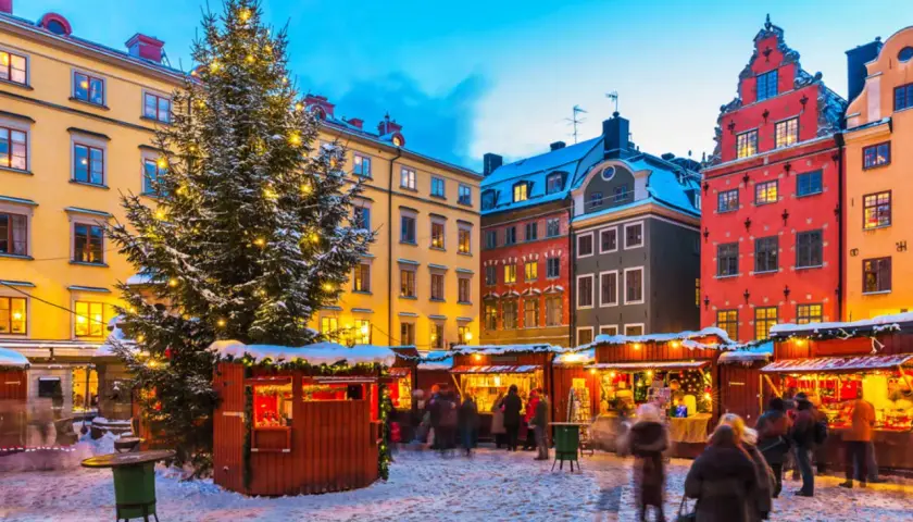 En snötäckt vinterviken julmarknad med ett stort, upplyst träd och färgglada hus. Folk strosar bland bodarna.