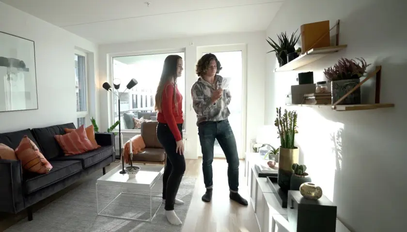 Två personer diskuterar olika inredningsstilar i ett ljust vardagsrum med modern möblering, en mörk soffa och hyllor med växter.