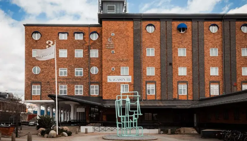 Magasin III, en byggnad i tegel med många fönster, har en flagga med texten "Magasin III". Framför finns en grön skulptur.
