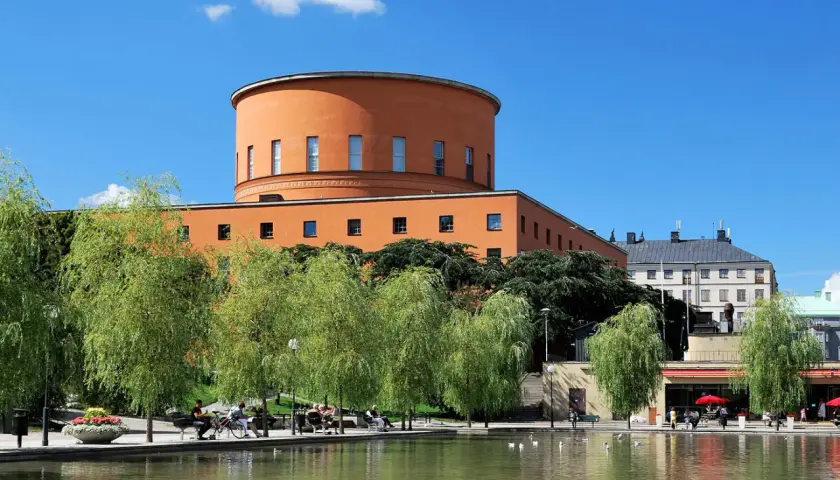 En vacker stadsvandring i Stockholm på egen hand. En orange, rund byggnad reser sig bakom grönskande träd vid en damm.