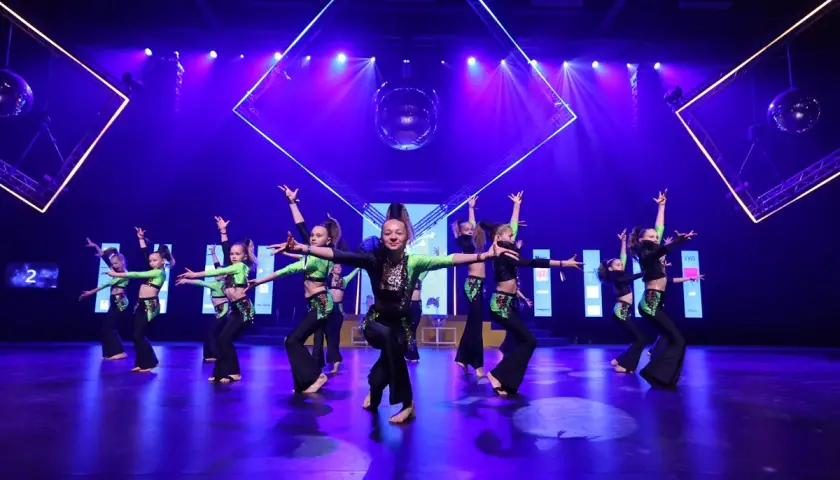 Ungdomar utför en energisk dansuppvisning i Stockholm, klädda i glittriga, gröna och svarta dräkter. Scenen är upplyst av blått och lila ljus, med en discokula som hänger ovanför.