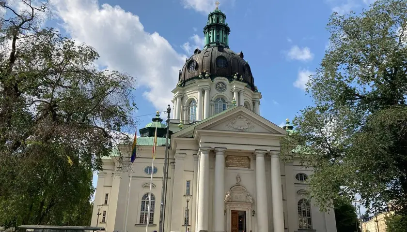 Imponerande Gustav Vasa kyrka med sin kupol och pelare, omgiven av träd och en blå himmel med moln.