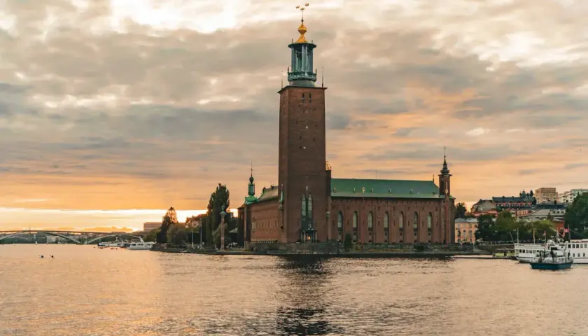 Stockholm Stadshus vid solnedgången, en vy som kunde vara från stockholm 1900. Vatten, båtar och en bro i bakgrunden.