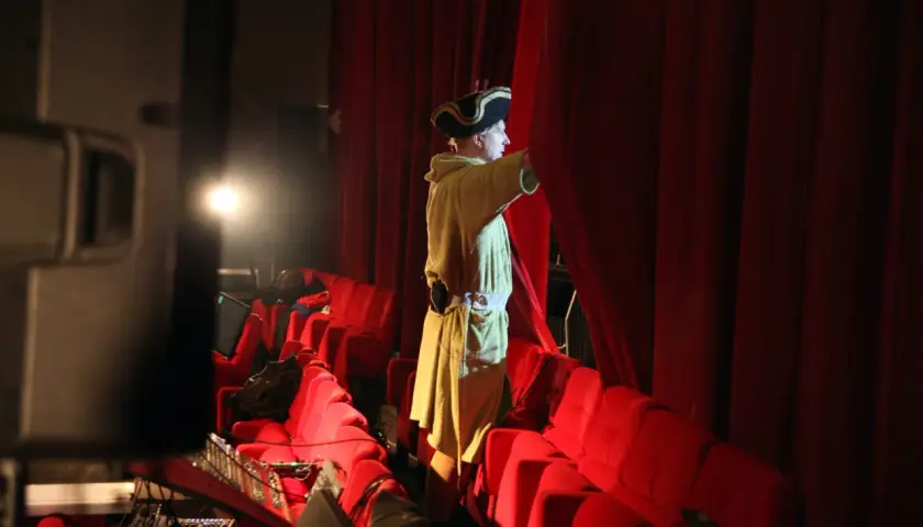 En person i historisk dräkt drar undan en röd ridå på en teater.
