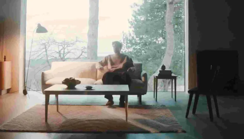 En man sitter i en soffa i ett ljust rum med stora fönster. En dansk designer kan ha skapat den moderna inredningen.