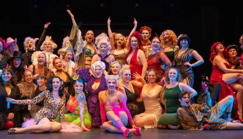 En stor grupp artister i glittrande kostymer poserar glatt på scenen under Stockholm Burlesque Festival.