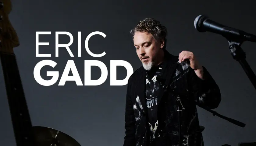 Eric Gadd, med sitt karaktäristiska lockiga hår och skägg, står redo att uppträda. En gitarr och mikrofon antyder en musikalisk kväll på Mosebacke.
