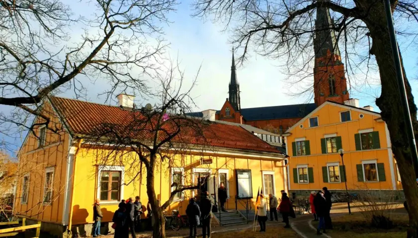 Gula hus med tegeltak och Uppsala domkyrka i bakgrunden. En grupp människor samlas utanför, kanske för ett besök på litteraturens hus Uppsala.