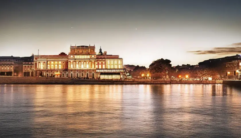 Kungliga operan i Stockholm lyser upp vid vattnet i skymningen, dess fasad speglas i det lugna vattnet.