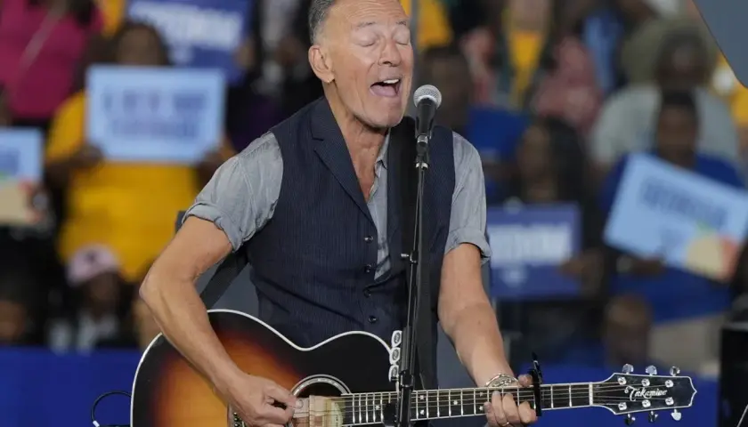 Bruce Springsteen sjunger och spelar gitarr på scenen. Publiken håller upp skyltar, vilket antyder ett kommande evenemang med Bruce Springsteen.