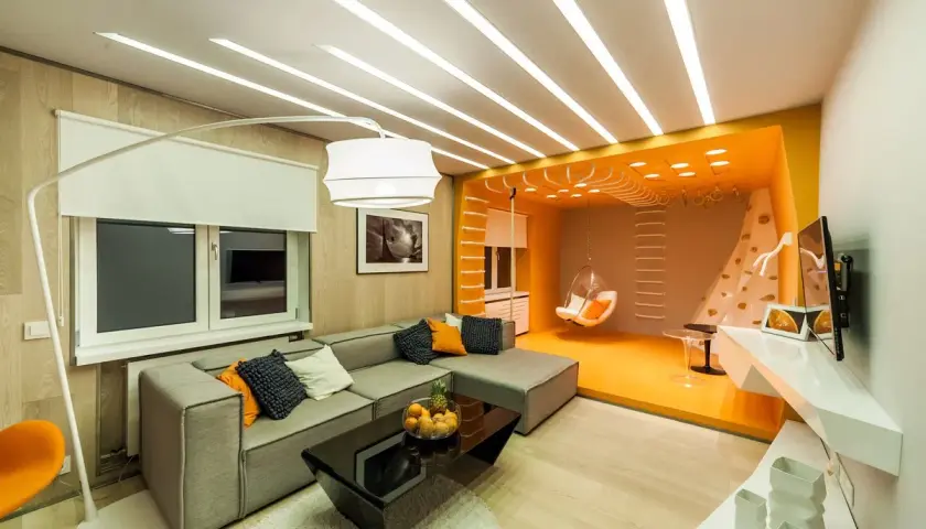Stort vardagsrum med en grå soffa, svart soffbord och en orange sittgunga. Taket har en unik design med många ljuslister.