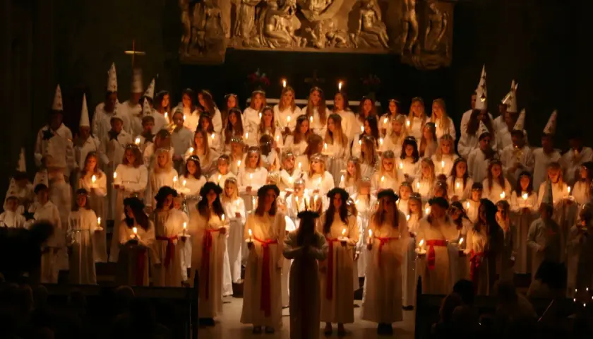 Stor grupp barn i vita dräkter och ljuskransar deltar i ett lucia firande i Stockholm.
