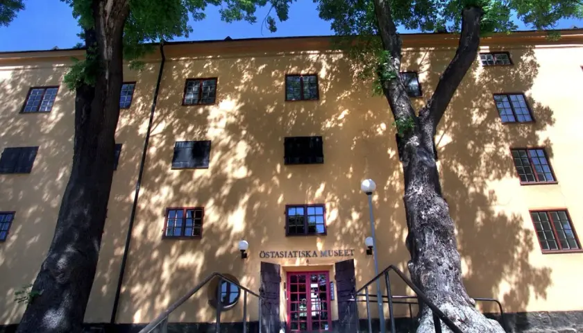 Östasiatiska museet i Stockholm, en gul byggnad med många fönster, omgiven av träd vars skuggor dansar på fasaden.