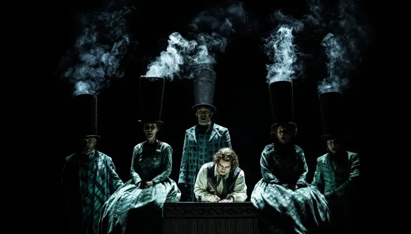 Scen från Sweeney Todd operan. Människor med höga hattar och rök som stiger ur dem. En man skriver vid ett bord.