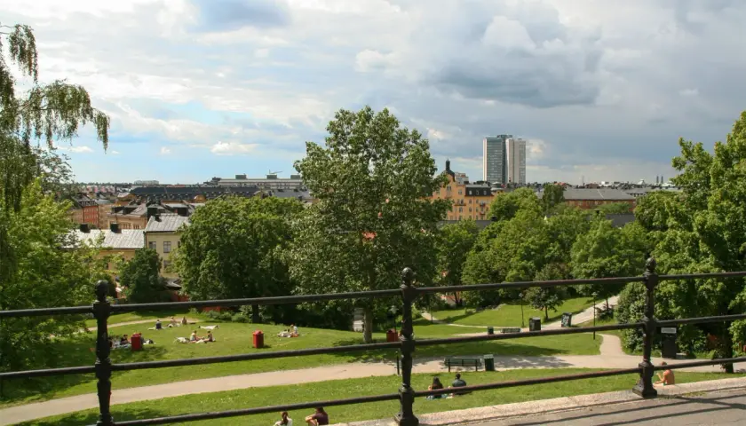 Sommar i Vitabergsparken, Stockholm. Människor njuter av solen på gräsmattan, med stadens takåsar och en teater i bakgrunden.