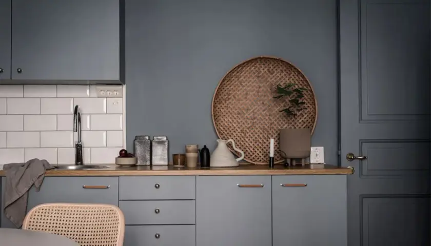 Ett kök med gråa skåp och en träbänk. En rund flätad bricka med en växt, en vit kanna och två metallburkar skapar ett vackert stilleben inredning.