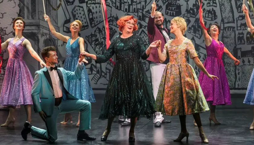 Scen från "Tootsie" på Oscarsteatern. En man i ljusblå kostym knäböjer medan en kvinna i glittrig grön klänning och andra i färgglada klänningar dansar.