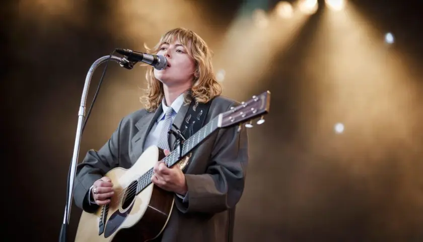 Ellen Krauss konsert. En artist med blont hår, klädd i kavaj och slips, spelar gitarr och sjunger i mikrofonen på en scen med strålkastare.