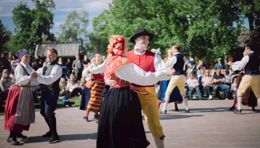 Dansande par i folkdräkter på en utescen, publiken tittar på. En härlig konsert på Skansen.