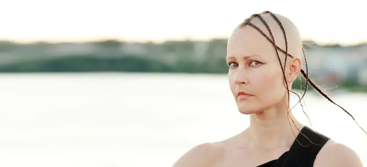 Virpi Pähkinen med flätor på rakad skalle, blickar mot betraktaren vid en sjö.