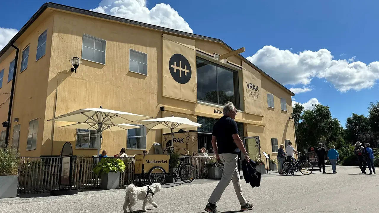 En gul byggnad med texten "VRAK MUSEUM OF WRECKS" under en stor svart symbol. Folk promenerar förbi, en hund går vid sidan av en man.