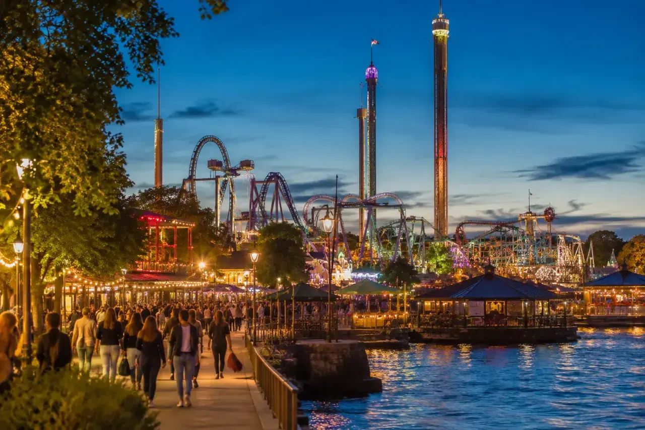 Kväll vid Gröna Lund med folk som promenerar längs vattnet, berg-och-dalbanor och höga torn som lyser upp himlen.