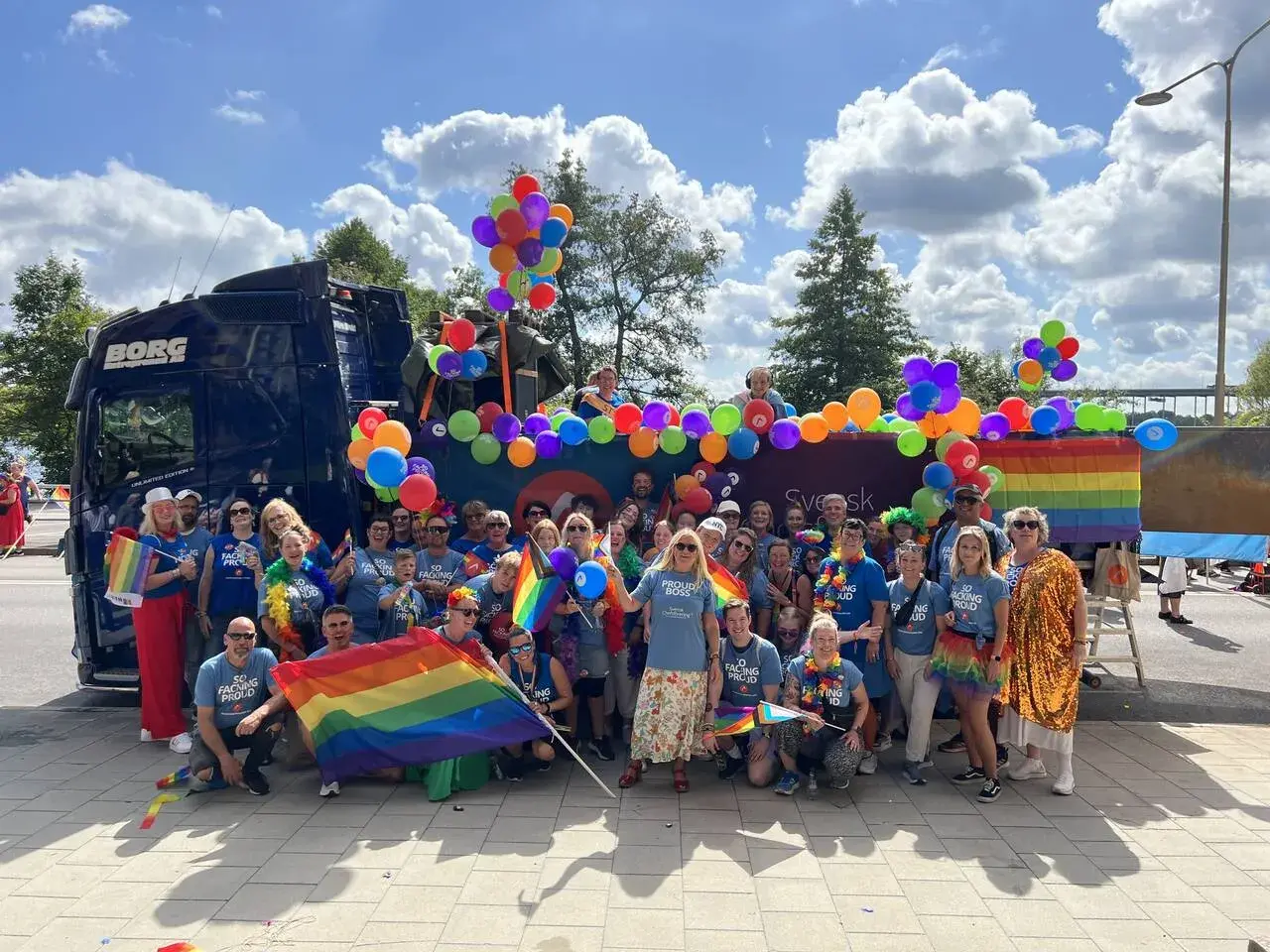 Stor grupp människor firar med regnbågsflaggor och ballonger vid en pride park.