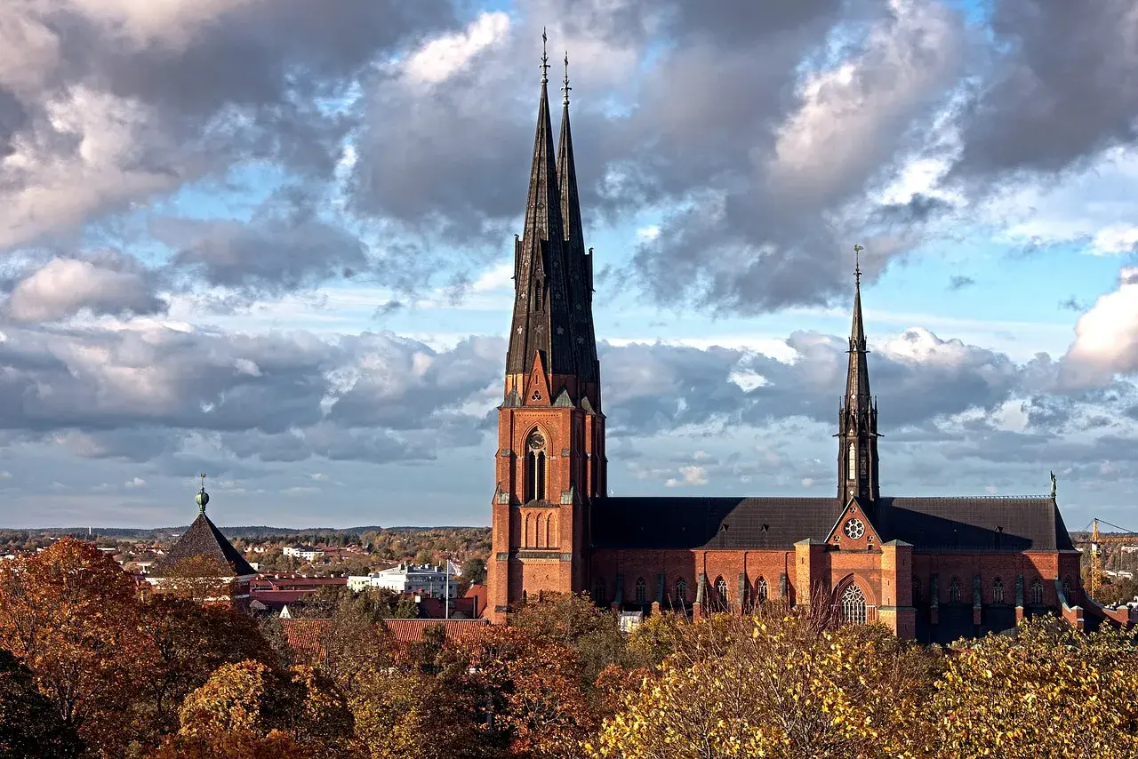 Uppsala domkyrka med sina höga torn reser sig mot en dramatisk molnhimmel. Höstlöven lyser i gult och orange.