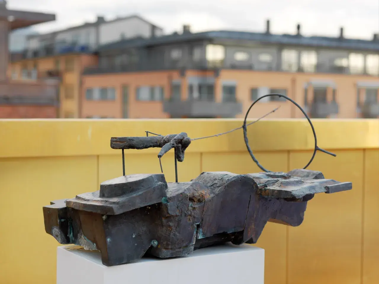 En abstrakt skulptur i mörk metall, placerad på ett vitt podium. I bakgrunden syns byggnader och en gul vägg, kanske vid museum Vasaparken.