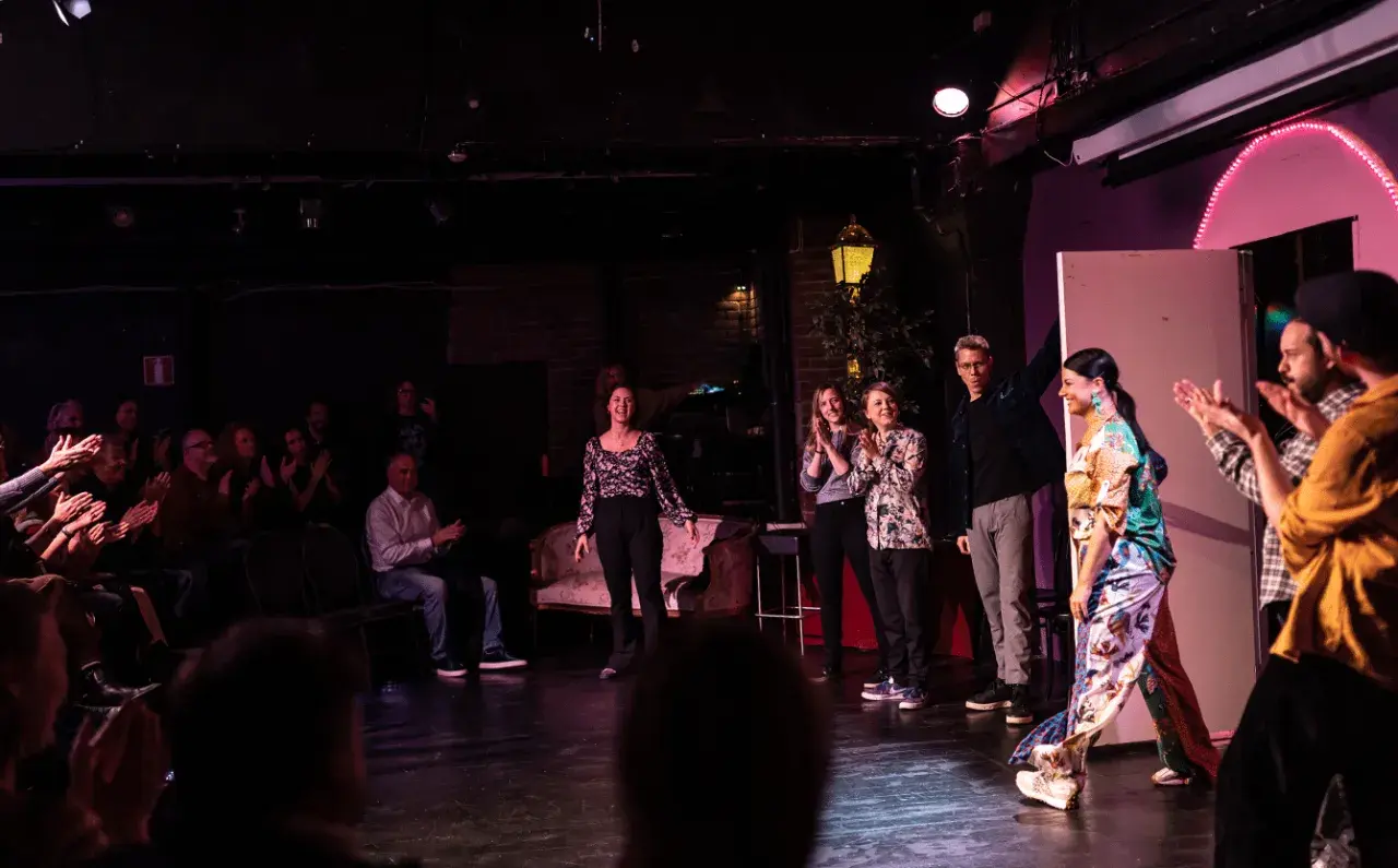 Publik applåderar en föreställning av improvisationsteater i Stockholm. Skådespelare på scenen tar emot hyllningar.