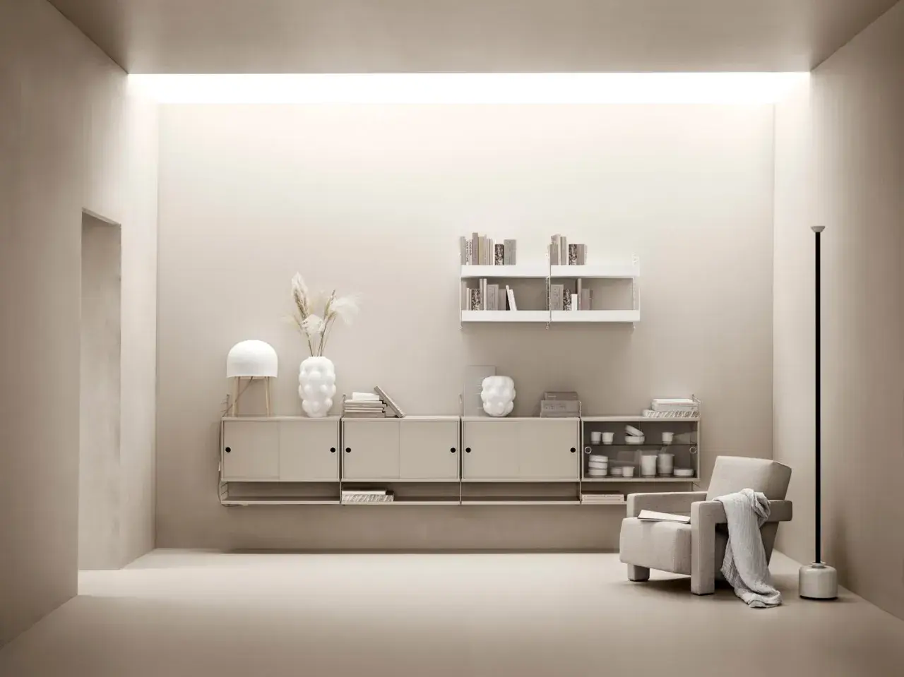 Minimalistiskt vardagsrum med stringhylla, beigefärgad förvaring, fåtölj och lampa.