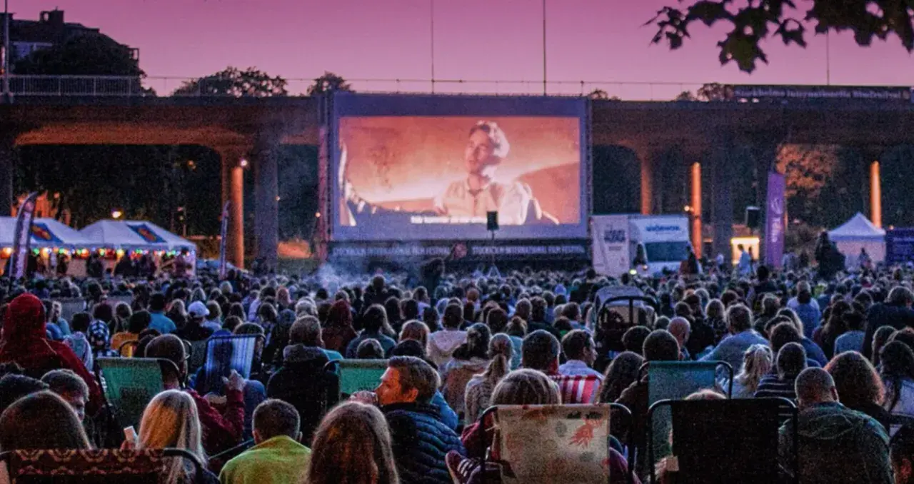 Stor publik njuter av sommarbio Stockholm under en lila kvällshimmel. En film visas på en stor duk framför tält och träd.