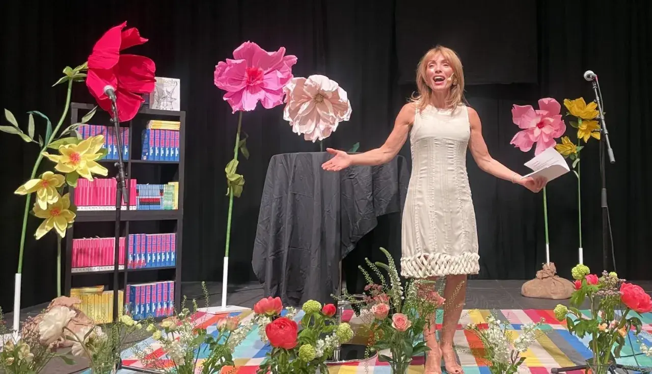 En kvinna talar på en scen med stora pappersblommor och bokhyllor, en del av Rinkeby bokfestival.