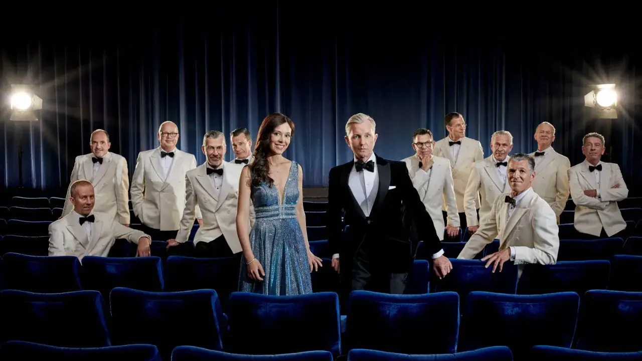 Max Raabe och hans Palast Orchester poserar i vita kostymer och en glittrig klänning framför en mörkblå ridå, redo för en föreställning i Stockholm.