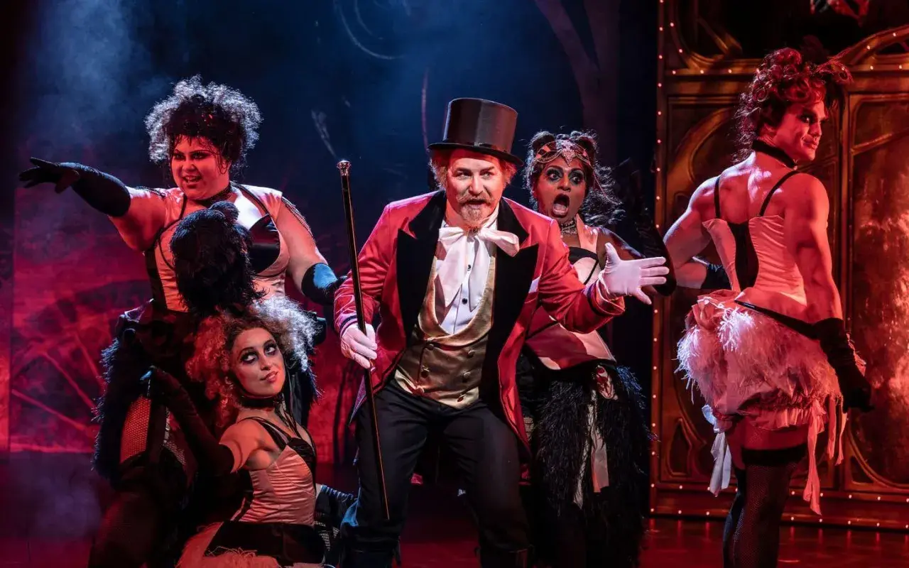 Scenen från Moulin Rouge! visar en cirkusdirektör med en cylinderhatt och en gyllene väst, omgiven av artister i glittrande kostymer. Handlar om kärlek och frihet i Paris.