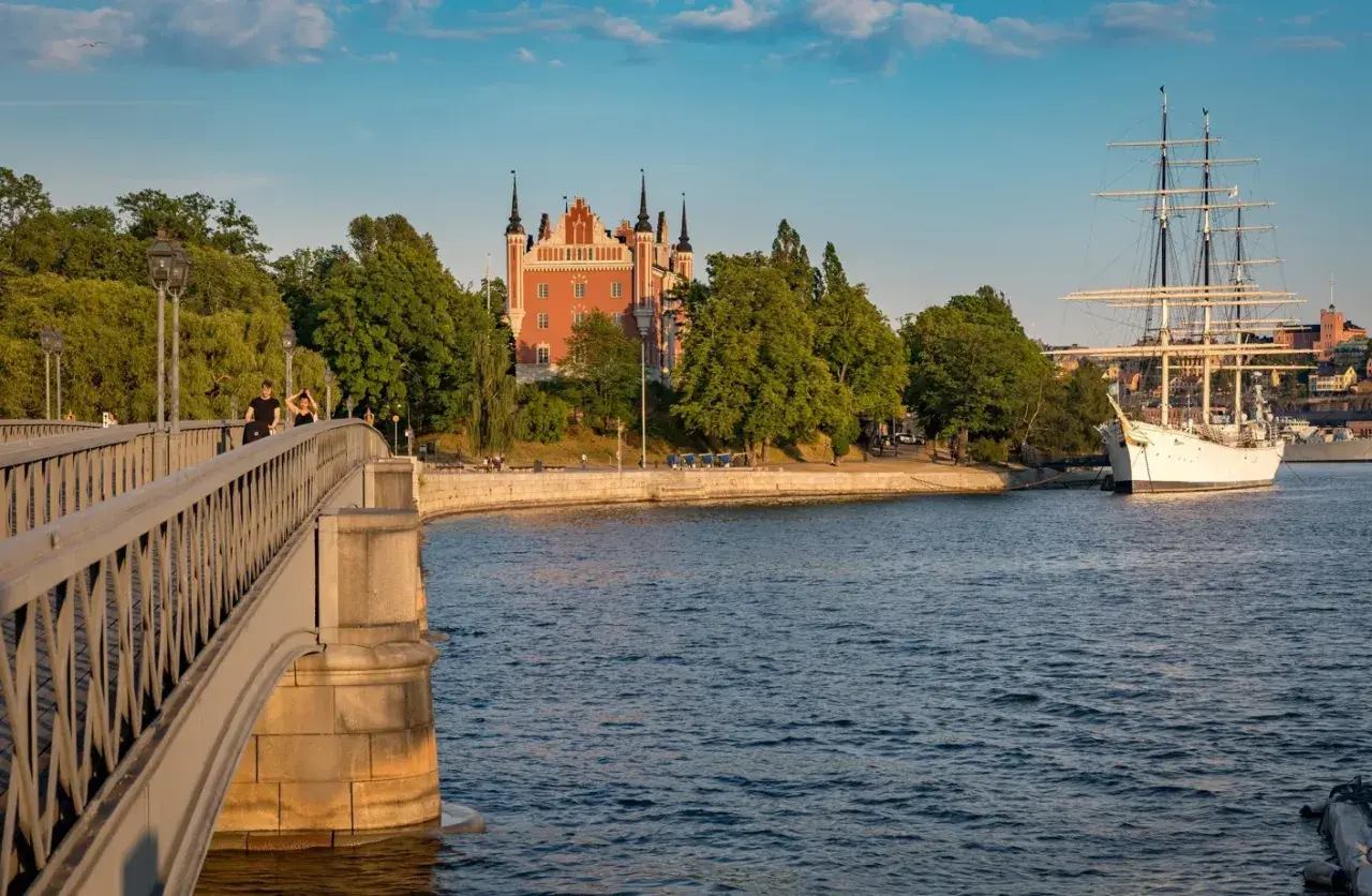 En vacker scen i Stockholm med en bro i förgrunden, ett rött tornliknande hus och ett stort vitt segelfartyg i vattnet.
