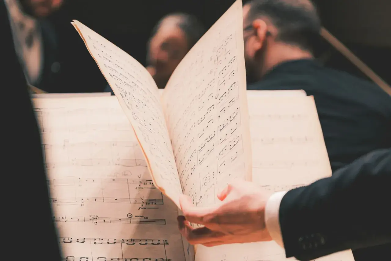 En hand bläddrar i noter under en lunchkonsert på operan. Musiker i bakgrunden förbereder sig.