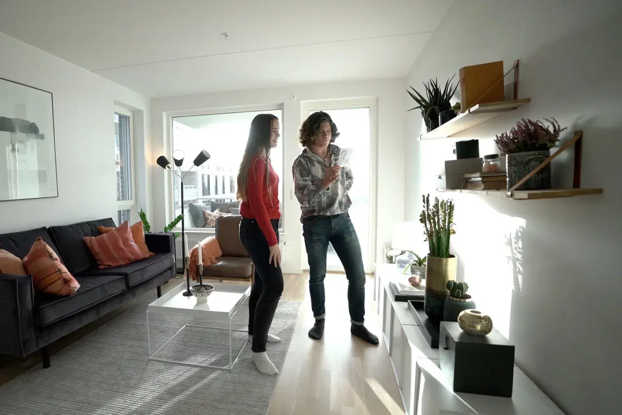 Två personer diskuterar olika inredningsstilar i ett ljust vardagsrum med modern möblering, en mörk soffa och hyllor med växter.
