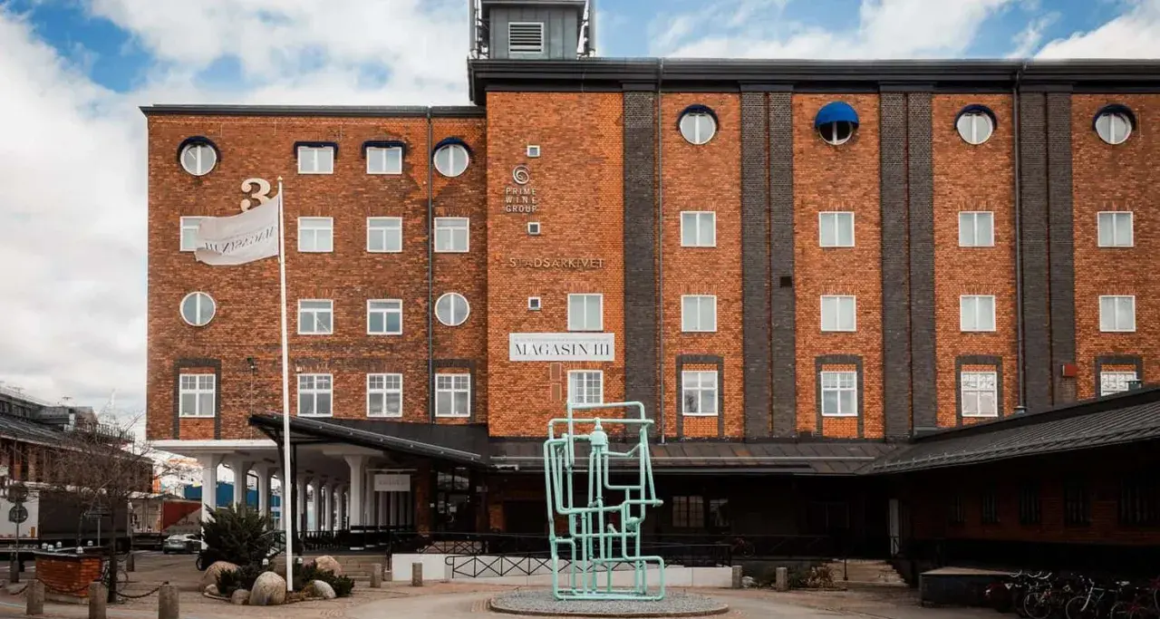 Magasin III, en byggnad i tegel med många fönster, har en flagga med texten "Magasin III". Framför finns en grön skulptur.