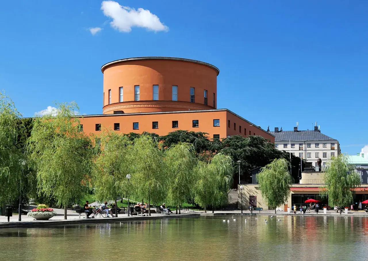 En vacker stadsvandring i Stockholm på egen hand. En orange, rund byggnad reser sig bakom grönskande träd vid en damm.