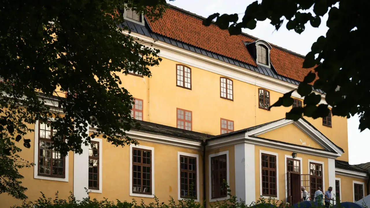 Gula fasaden på Ulriksdals teater med tegeltak och mörkgröna trädgrenar som ramar in.