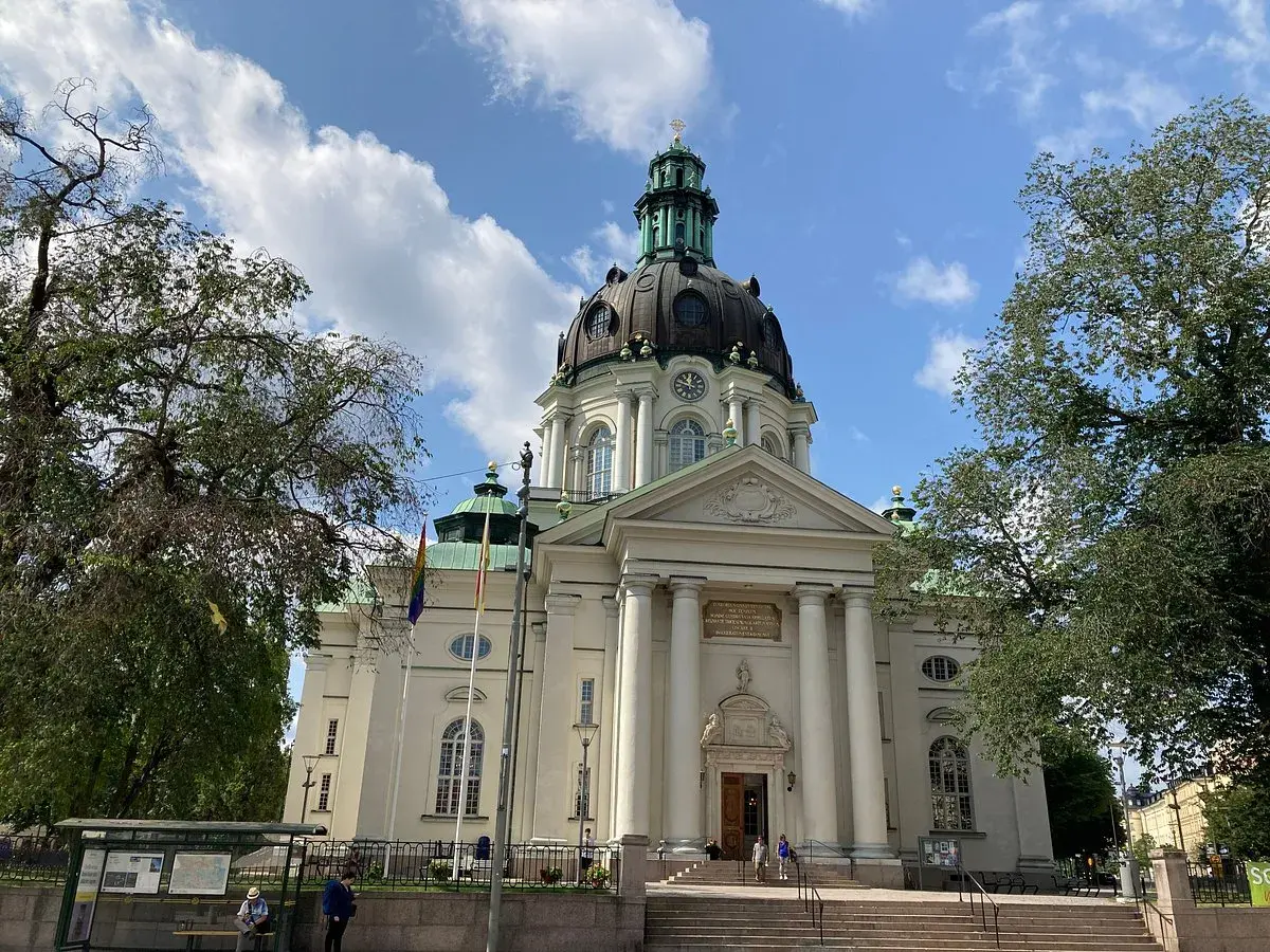 Imponerande Gustav Vasa kyrka med sin kupol och pelare, omgiven av träd och en blå himmel med moln.