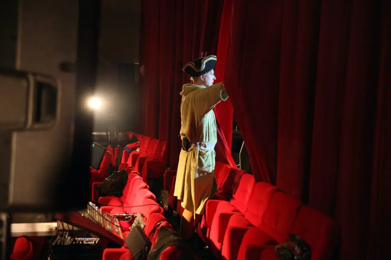 En person i historisk dräkt drar undan en röd ridå på en teater.