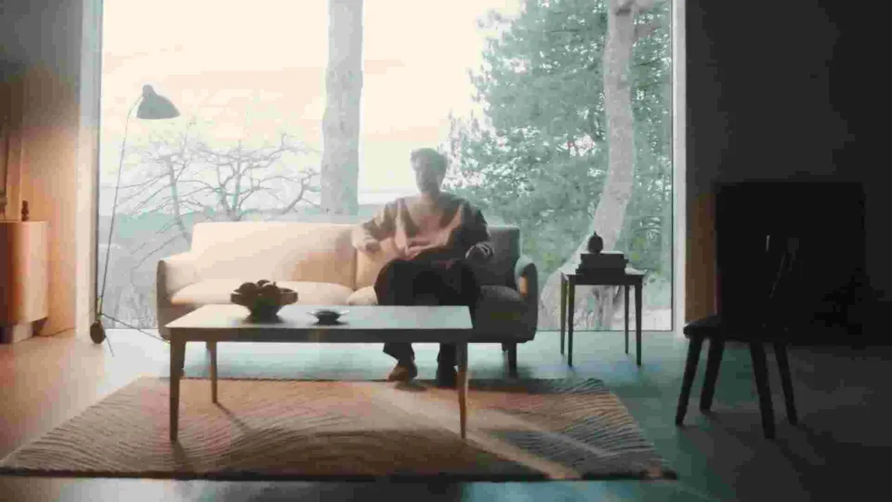 En man sitter i en soffa i ett ljust rum med stora fönster. En dansk designer kan ha skapat den moderna inredningen.