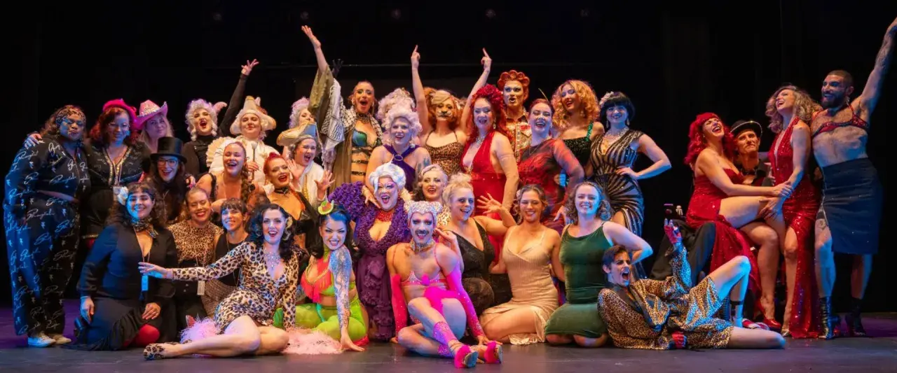 En stor grupp artister i glittrande kostymer poserar glatt på scenen under Stockholm Burlesque Festival.