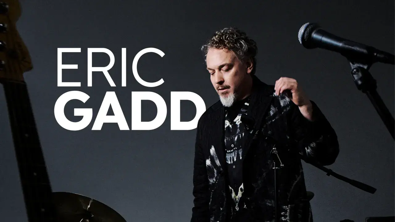 Eric Gadd, med sitt karaktäristiska lockiga hår och skägg, står redo att uppträda. En gitarr och mikrofon antyder en musikalisk kväll på Mosebacke.
