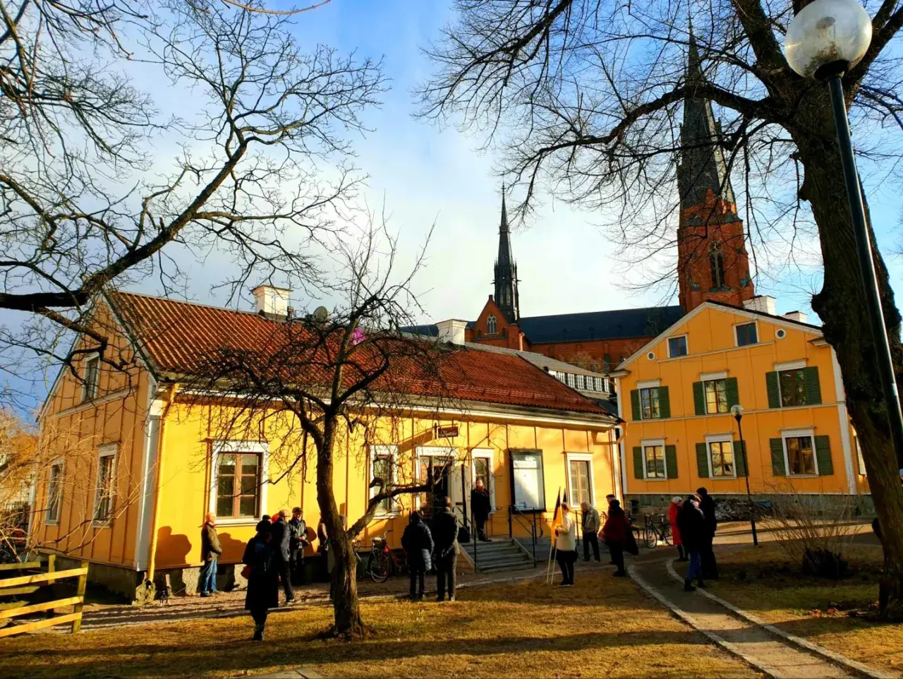 Gula hus med tegeltak och Uppsala domkyrka i bakgrunden. En grupp människor samlas utanför, kanske för ett besök på litteraturens hus Uppsala.